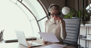 Young business woman secretary working alone in modern office. Female receptionist wearing headset using laptop, checking papers at workplace. Professional manager drinking water sitting at desk.