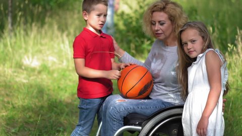 Mom in a wheelchair for a walk with her family and communicates with two children standing nearby holding her son ball on his feet and father standing in the distance. Family concept. Prores 422. 