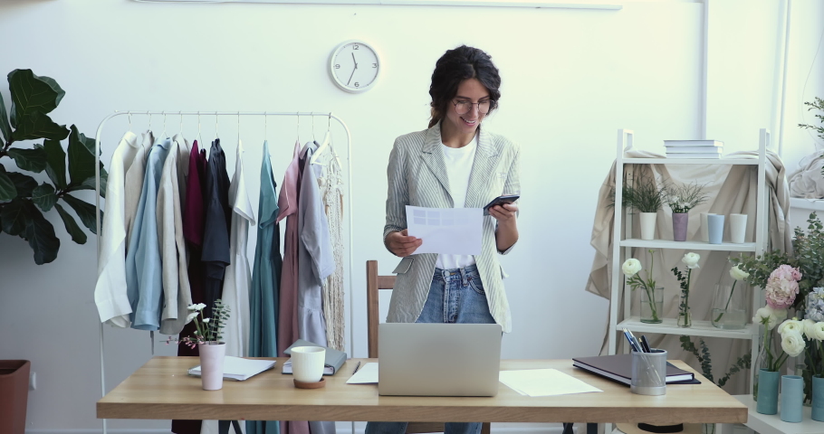Smiling young female fashion designer stylist holding swatches palette using smart phone at workplace, texting message, communicating with customer, taking clothing online atelier order in cell app.