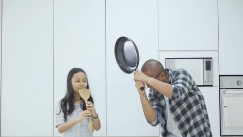 Happy little girl playing a spatula as a flute to her father like swayed snake while having fun in the kitchen. Shot in 4k resolution