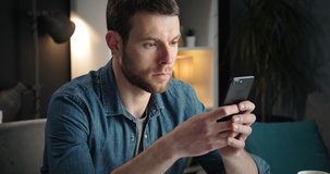 Close up of bearded man in denim shirt spending free time at home with smartphone. Young male with dark hair sitting on couch with digital device.
