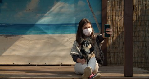 A teen age girl makes selfie while wearing protecting mask. She tries to make a photo with her dog. Self isolation in countryside while dreaming about sunny beach. Sunny backyard. : vidéo de stock