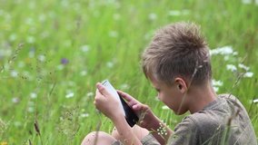 Closeup side view video footage of cute blonde white kid sitting in scenic green meadow in countryside and using modern black smartphone.