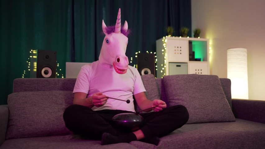 a man in a mask unicorn sits at home and plays around on the hapi drum warm atmosphere on isolation