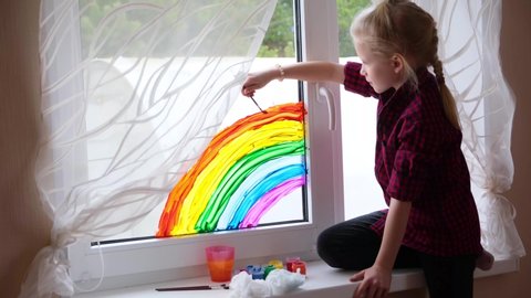 4k. Girl painting rainbow on window during Covid-19 quarantine at home. Stay at home social media campaign for coronavirus prevention, let's all be well, hope. Chase the rainbow.
