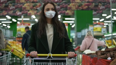 Young woman wearing protective medical face mask rolls shopping cart at grocery store. Female customer picks vegetables at economy supermarket during covid-19 coronavirus pandemic, social distancing