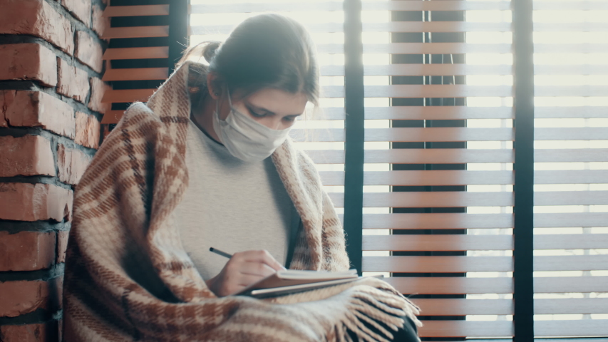 Young female teenager in protective medical mask during pandemic covid-19, she is sitting on windowsill in loft design house and writes something in notebook, brick walls, windows with jalousie