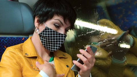 Woman in protective mask on face uses smartphone, sitting by window of train. Communication during social distance in public transport. Surprise reaction of reading covid-19 pandemic breaking news.