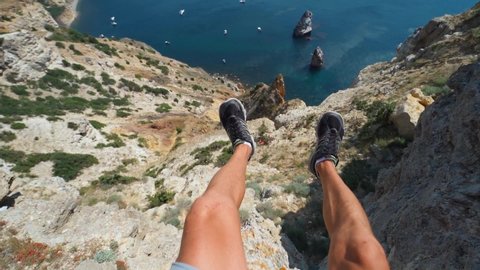 POV first person view male legs in black sneackers sitting on cliff edge with amazing view seascape with high limestone cliff over blue sea and yachts.Traveler enjoying recreation travel