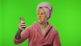 Portrait of young man wearing pink bathrobe and face cleaning mask after shower doing beauty procedures and using smartphone social network app for video call. Funny-looking. Chroma key