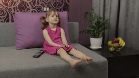 Cute little kid sitting on couch while watching TV funny music movie at home in the evening. Happy young child girl watch television on living room. Concept video game, entertainment, family, relax