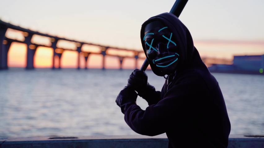 Man in the glowing blue mask threatens waving a bat in hand. At background sunset and the bridge at the lake | Shutterstock HD Video #1052318845