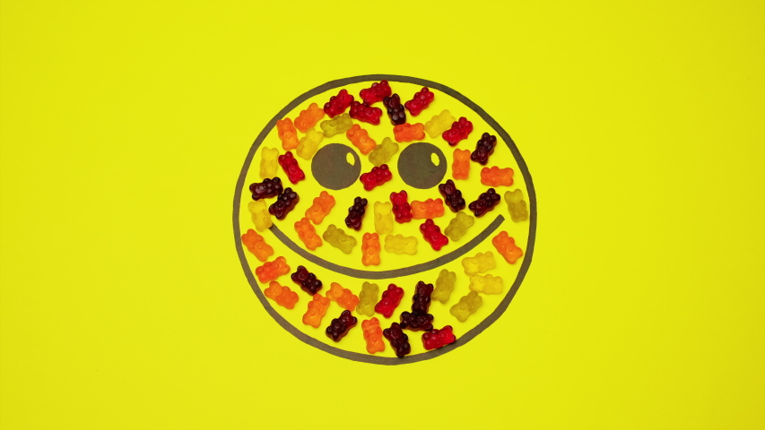 Smiley On Yellow Background Filled With Colored Candies. Candies In The Form Of Bears Move From The Smiley. Then Come Back.