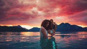 Dreamy Cinemagraph / seamless video loop of a beautiful young model girl standing in the calm waves of lake Walchensee with an old golden mirror in Germany/Bavaria in a white fashion dress by sunset.