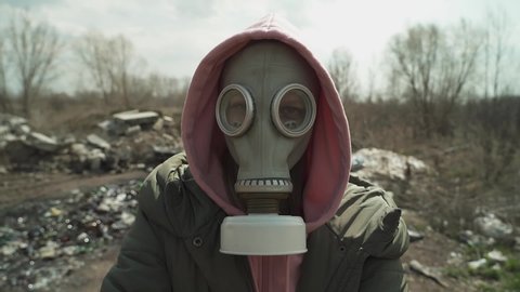 The girl in the gasmask in a pink hoodie and looking at camera on the background of the landfill. mobile camera.