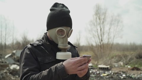 Man in the gasmask and a black hat. amid the landfill. swipe smartphone.