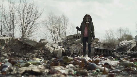 The girl in the gasmask walks amid the landfill. overall plan. looks around in search of food.