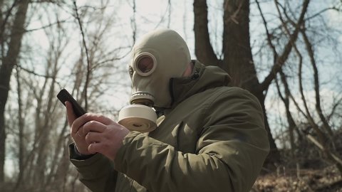 Man in gasmask and hood in the woods. the view of the profile. looking smartphone.