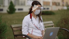 Young female doctor in plastic shield and surgical mask uses laptop outdoors