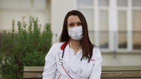 Medical online webinar concept: Young female doctor in surgical mask and earbuds looks at webcam outdoors