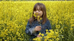 A beautiful little girl with posing against a yellow rapeseed field. She smelling fresh yellow canola flowers. Slow motion video.