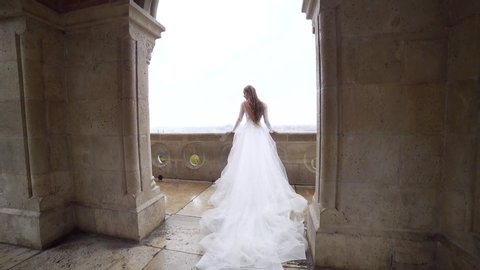 beautiful fairy princess overlooks the balcony or terrace of an exquisite ancient castle against the backdrop of the old town and river. Bride in white wedding dress. Landscape. Slow motion. behind	