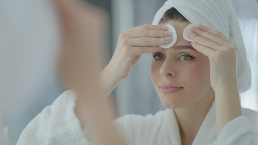 Beautiful woman in white towel on her head and a bathrobe looking in reflection in the mirror and cleansing face skin removing make up with cotton disk, skincare concept.
 | Shutterstock HD Video #1052346790