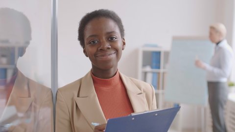 Medium shot portrait of young African woman standing in office with clipboard in her hands and looking at camera while her colleague working on background
