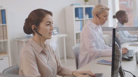 Medium shot of three multiethnic female operators wearing white shirts and headphones sitting at computer displays in the office and consulting interlocutors by phone