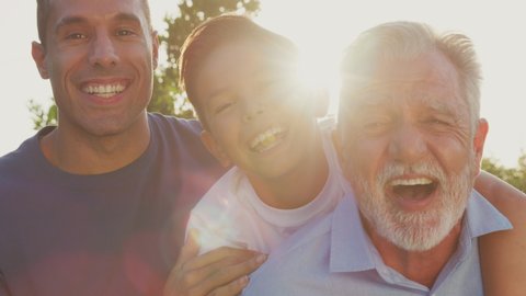 Multi-generation male Hispanic family relaxing in garden at home together with grandfather giving grandson piggyback against flaring sun - shot in slow motion