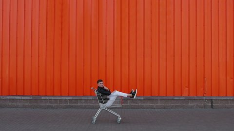 Happy customer man is sitting in a shopping cart and rides along the wall of mall store center outside and imitates smoking. Discounts and sales concept. Funny and amusement moment