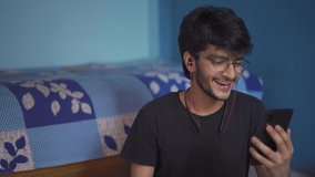 A happy young Indian boy using smartphone and wireless headphones to video call with friends in the lock down period. Stay at home. 4k 