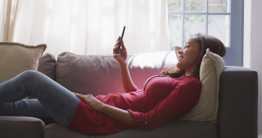 Mixed race woman enjoying her time at home, lying on a couch, using a smartphone and smiling, social distancing and self isolation in quarantine lockdown, in slow motion