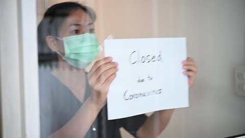 Asian business owner woman wearing face mask or small shop manager attaching business closed sign at shop entrance due to financial crisis from coronavirus covid-19 epidemic outbreak over the world.