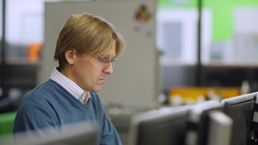 Tracking side view shot of focused middle aged engineer in eyeglasses working on computer sitting at desk in laboratory