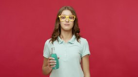 Joyful young woman with bright eyeglasses holding blue cocktail and dancing, drinking alcohol at party, celebrating weekend, looking carefree optimistic. indoor studio shot isolated on red background
