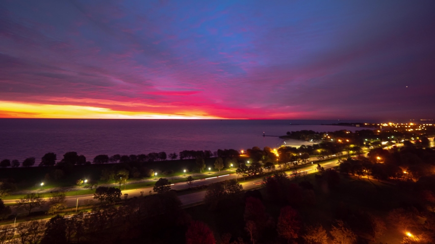 A beautiful panning up aerial sunrise timelapse with sky on fire pink, yellow, blue, purple, orange and red eruption of color on the clouds as traffic builds on Lake Shore Drive below in Chicago.