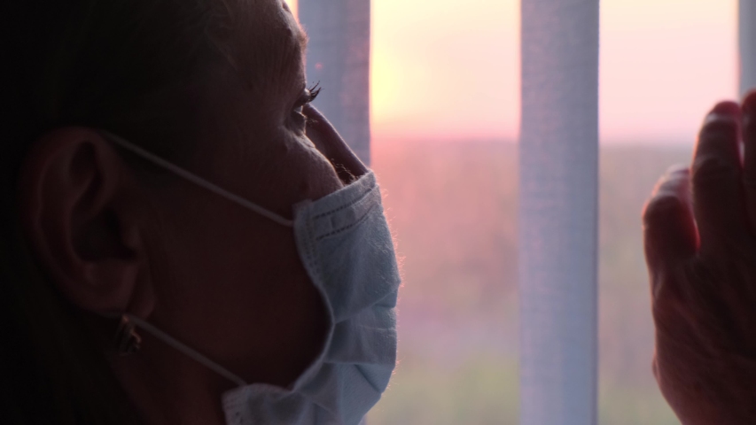 An elderly woman in a protective mask by the window at sunset. Doctor in a protective mask looks out the window at sunset | Shutterstock HD Video #1052415490