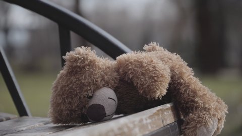 Close-up of teddy bear lying on wooden bench in city park. Lost toy outdoors. Concept of loneliness, abandonment, kidnapping, lost kids.