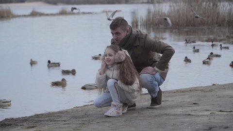 Cheerful Caucasian man and girl watching at ducks and imitating fly. Portrait of happy young father and pretty daughter enjoying weekends outdoors. Happiness, family, joy, leisure.
