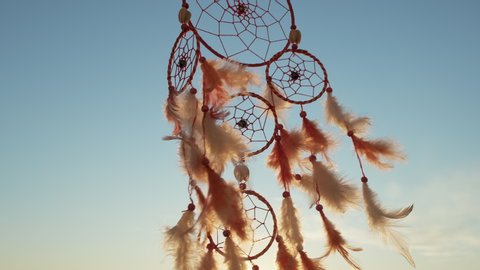 Dreamcatcher with orange feathers rotate, swaying with light wind slow motion slide against blue sky at sunset in summer. Boho style sunset light on sunny day. Boho style amulet. Vintage. Macro