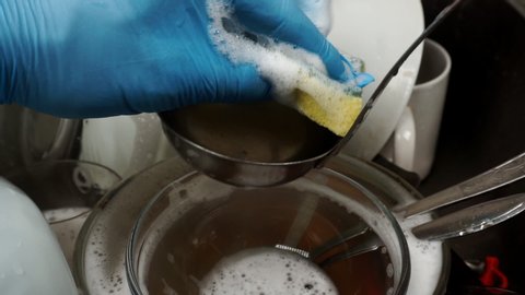 A male hand in a rubber glove washes dishes with a foamy sponge for washing dishes. In the background is a mountain of dirty dishes.