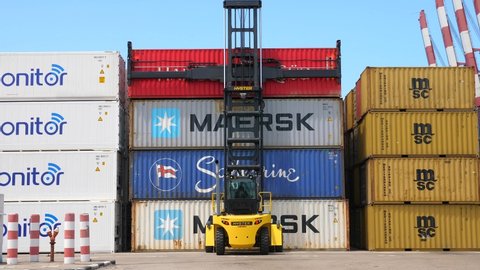 Haifa, Israel - May 11, 2020: Container handler lifting a Red Hamburg Sud Shipping container off a truck and stack it on a storage platform.