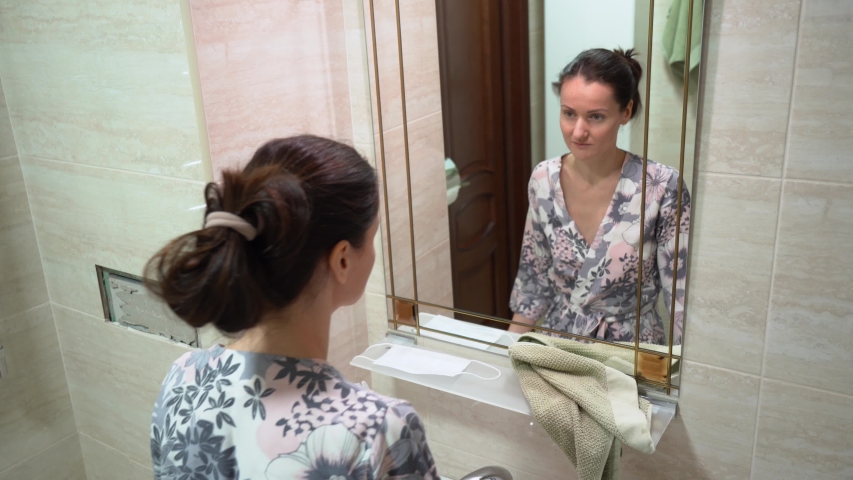 Young girl with hair bun in pajamas wash her face, wipe with a green towel, puts on protective medical mask, look at herself in front of the mirror in bathroom. Covid 19 virus protection concept.