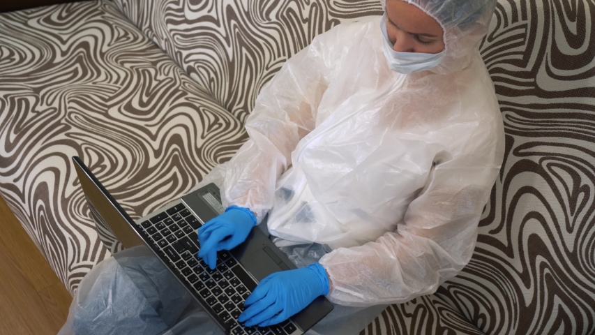 Caucasian woman wear protection white suit, medical face mask, blue rubber gloves, sits on couch, using laptop at home. Isolated girl typing text, working remote. Self-isolating. Lock down. Top view