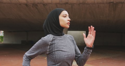 Side view of a mixed race woman wearing sportswear and hijab, exercising outdoors in the city on a sunny day, running in slow motion
