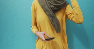 Front view of a mixed race woman wearing hijab in the city, leaning and using smartphone, wearing headphones and listening to music in slow motion