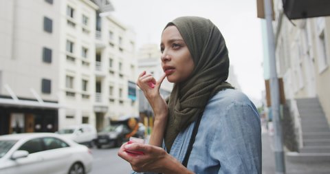 Side view of a mixed race woman wearing hijab in the city on a sunny day, standing and putting on lip balm, in slow motion