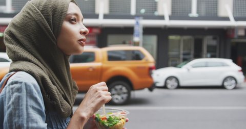 Side view close up of a mixed race woman wearing hijab in the city on a sunny day, walking in the street and eating a takeaway salad in slow motion
