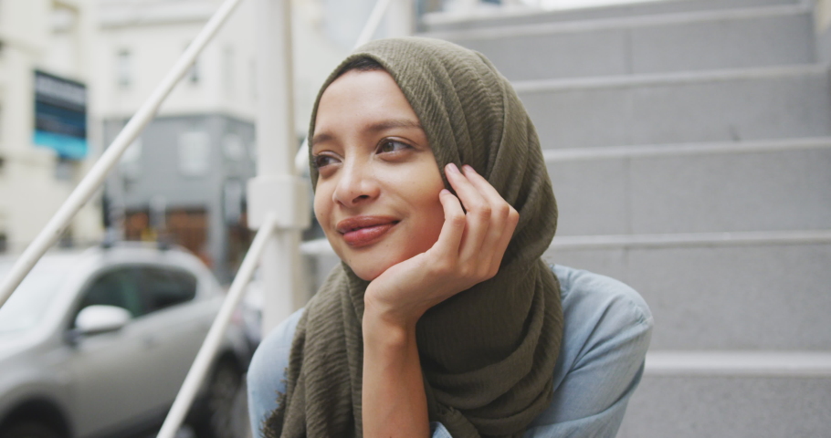 Front view close up of a mixed race woman wearing hijab in the city on a sunny day, sitting on stairs and thinking in slow motion